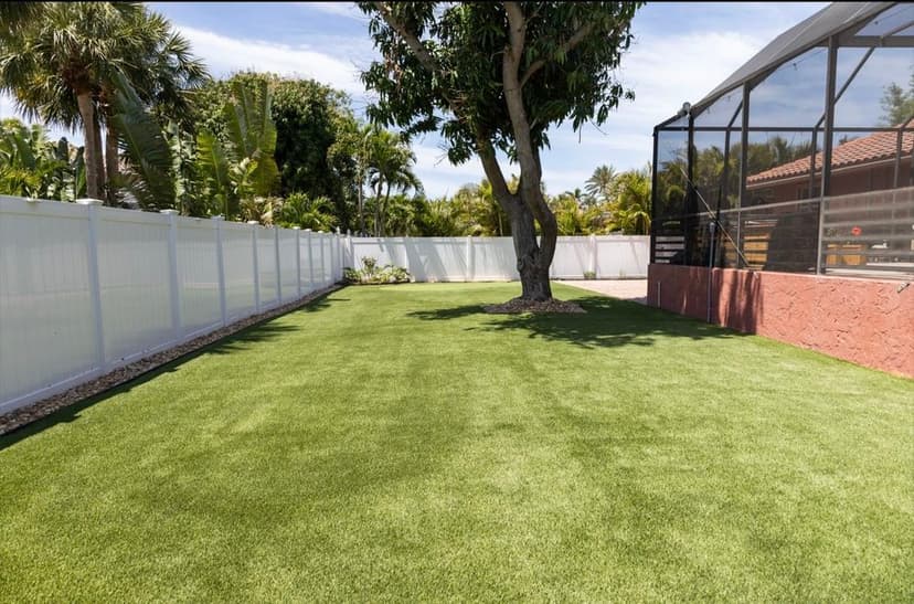 Lush green backyard with artificial turf, surrounded by a white fence and tropical landscaping.
