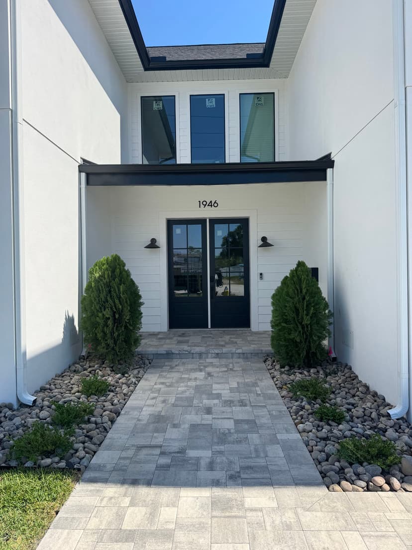 Modern entryway of a white house with stone pathway and lush greenery, featuring double doors.